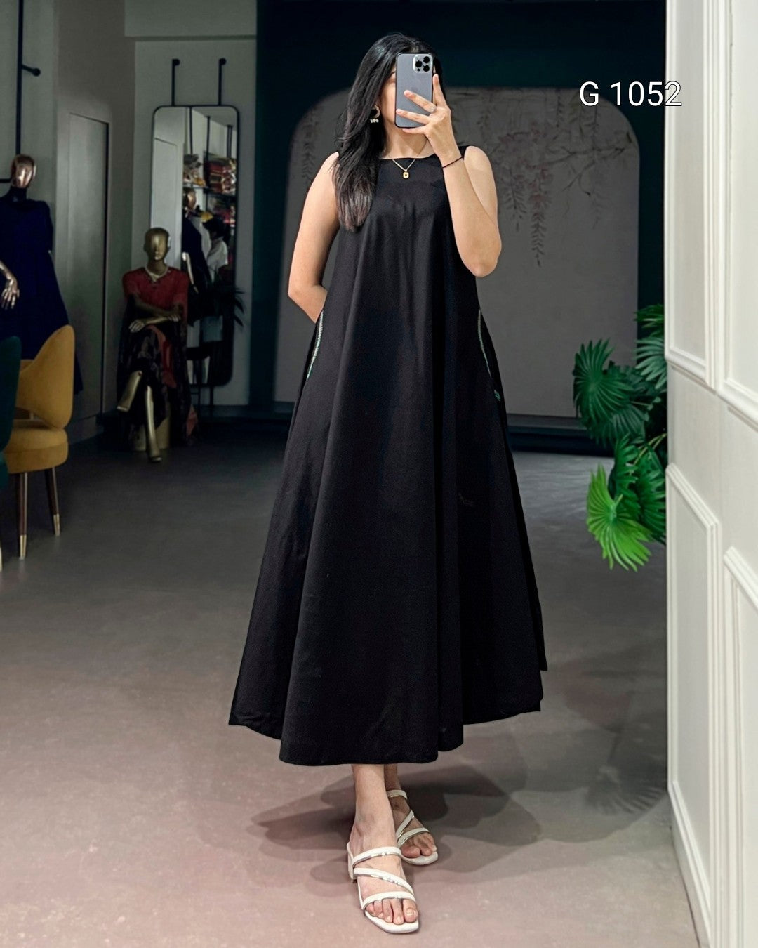 A woman takes a mirror selfie wearing a sleeveless black midi dress with a flared skirt and white strappy sandals. The setting is a boutique interior with mannequins, a yellow chair, and a potted plant visible in the background.