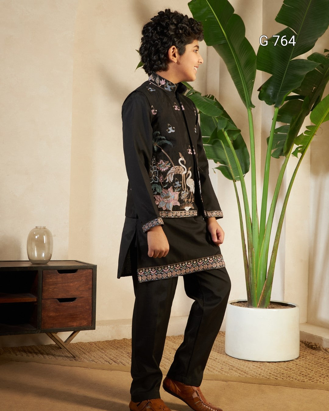Young boy in formal attire standing in a room with a plant and wooden cabinet.