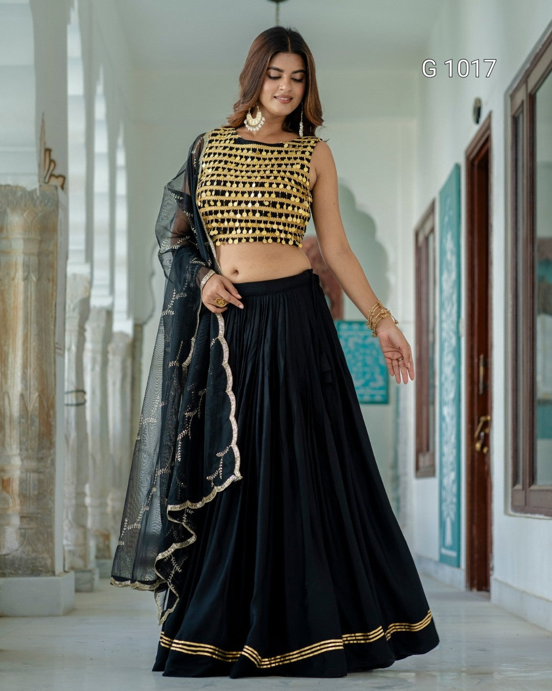 A woman models a black and gold traditional Indian outfit consisting of a black pleated skirt, a gold embroidered sleeveless top, and a black sheer dupatta with gold borders, set against a white corridor background.