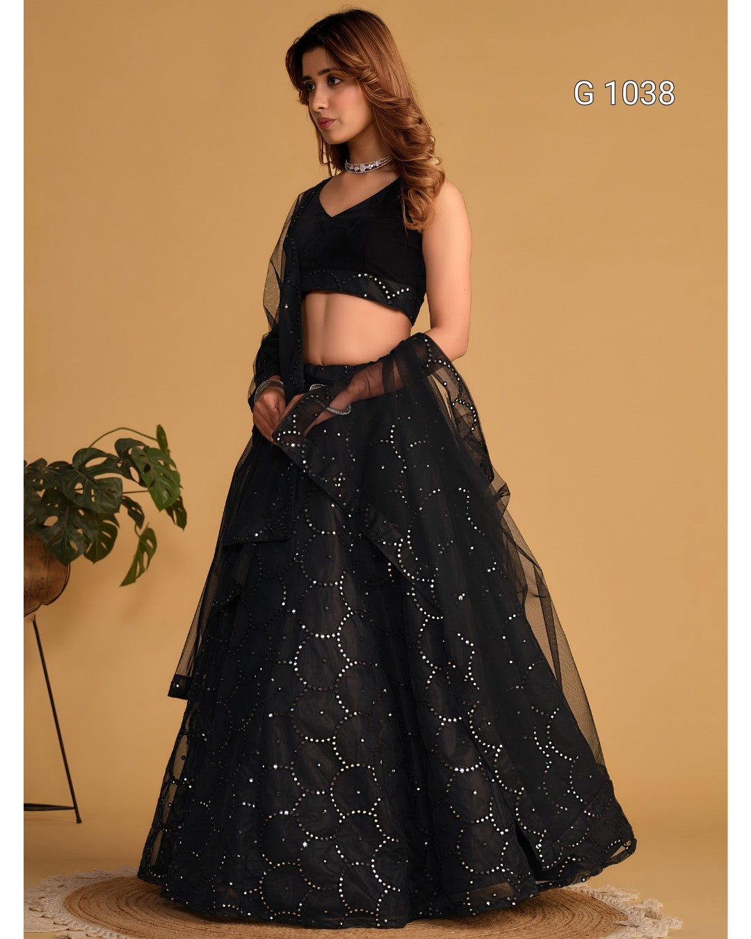 A woman modeling a black Indian ethnic wear set consisting of a sleeveless crop blouse, a long sheer sleeve, and a flared black netta lehenga skirt decorated with silver mirror work, set against a solid beige background.