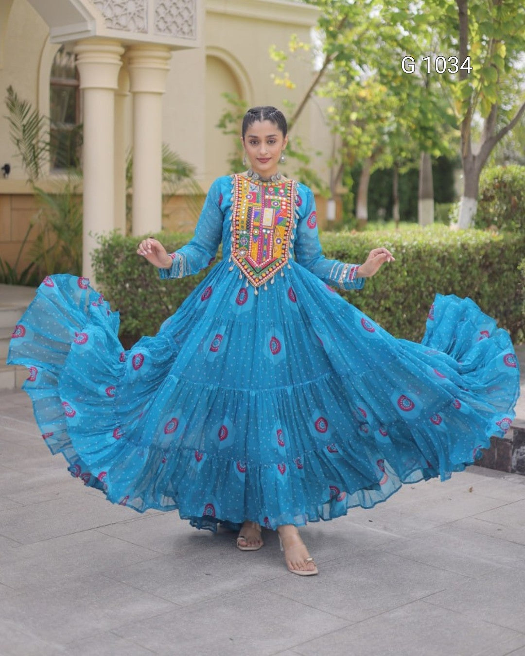 A woman wearing a bright blue tiered Anarkali gown with a heavily embroidered multicolor neckline, captured mid-spin as the dress flares out. The setting is an outdoor courtyard with beige pillars, green hedges, and a paved ground.