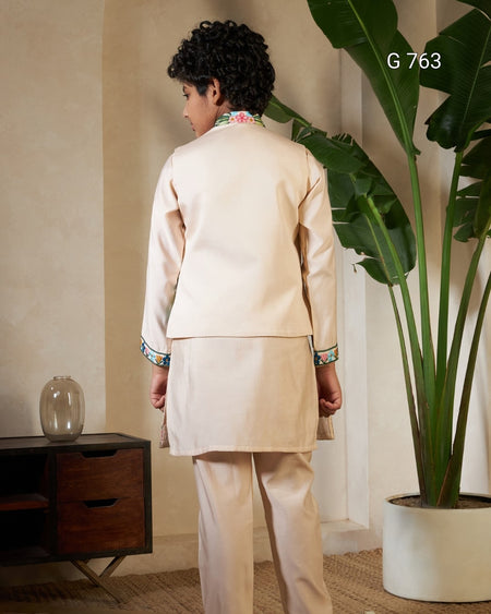 Person wearing a beige traditional outfit standing in a room with a plant and wooden cabinet.