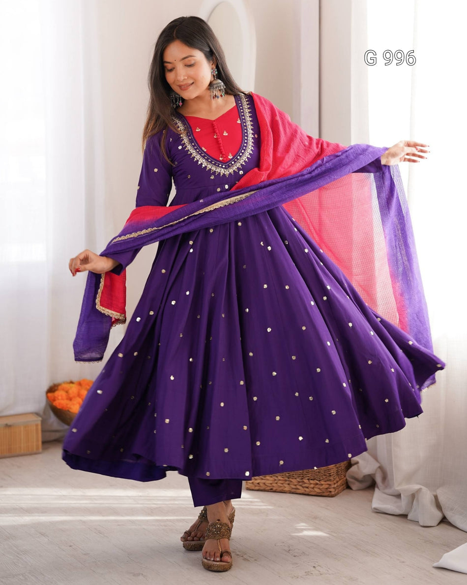A woman modeling a purple traditional Anarkali gown with a red embroidered neckline, gold dot embellishments, and a pink dupatta, standing in a bright room with white curtains.