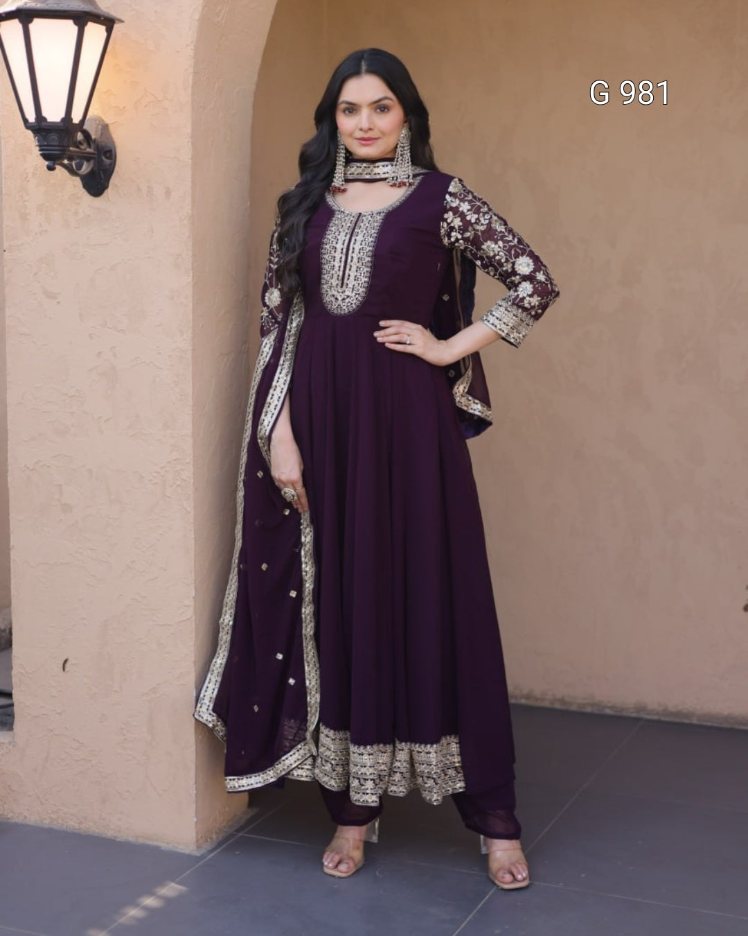 A woman models a deep purple Anarkali gown with ornate silver and gold embroidery, paired with a matching dupatta, set against a beige stucco wall background.