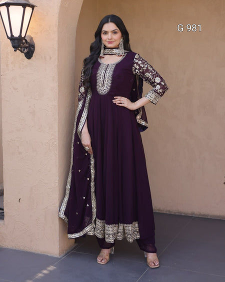 A woman models a deep purple Anarkali gown with ornate silver and gold embroidery, paired with a matching dupatta, set against a beige stucco wall background.