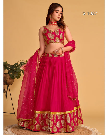 A woman models a bright pink lehenga choli set with gold leaf embroidery, featuring a matching dupatta, set against a solid beige background.