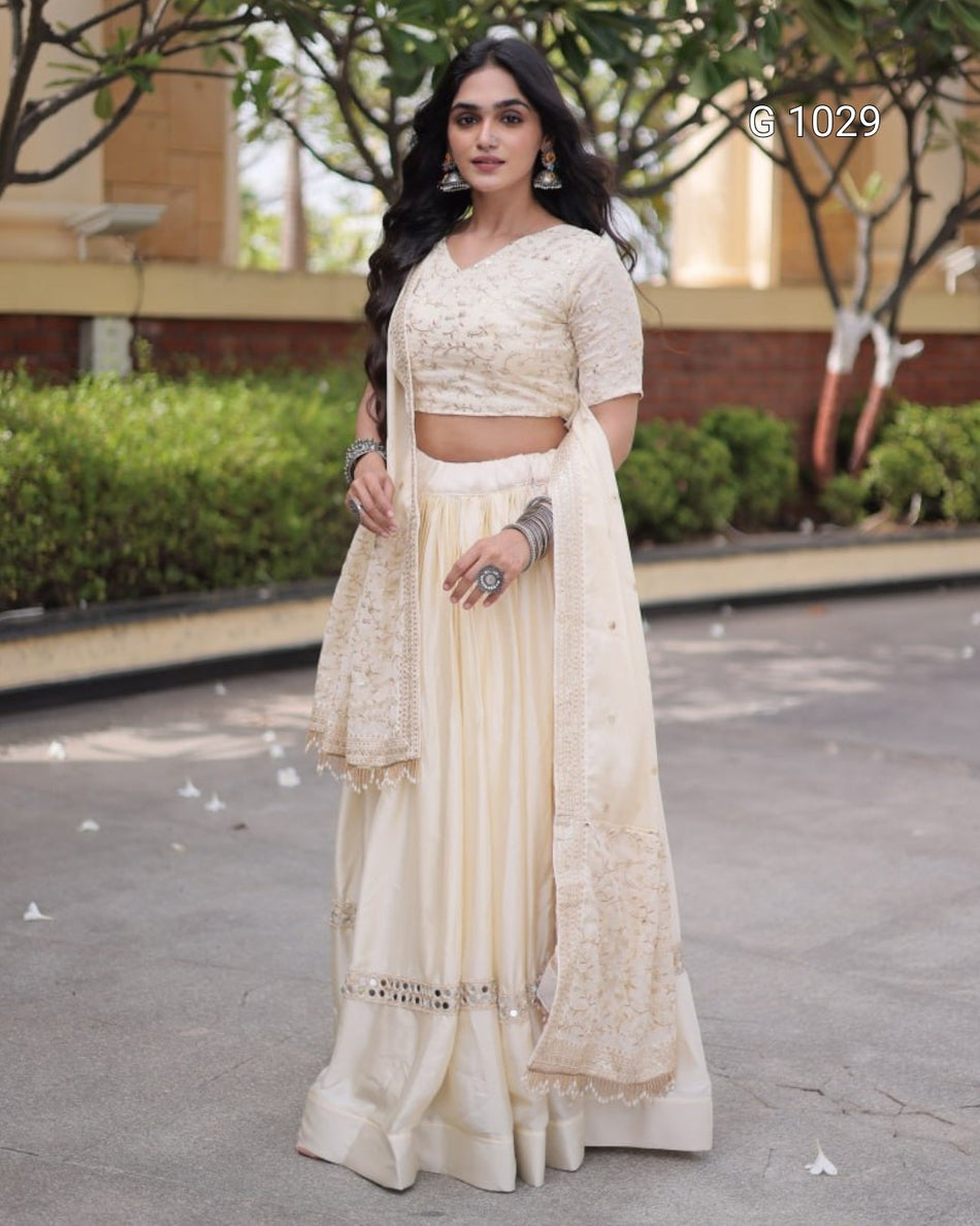 A woman modeling a cream-colored traditional Indian outfit consisting of a short-sleeved embroidered blouse, a matching flared skirt, and a draped dupatta, standing outdoors on a paved walkway.