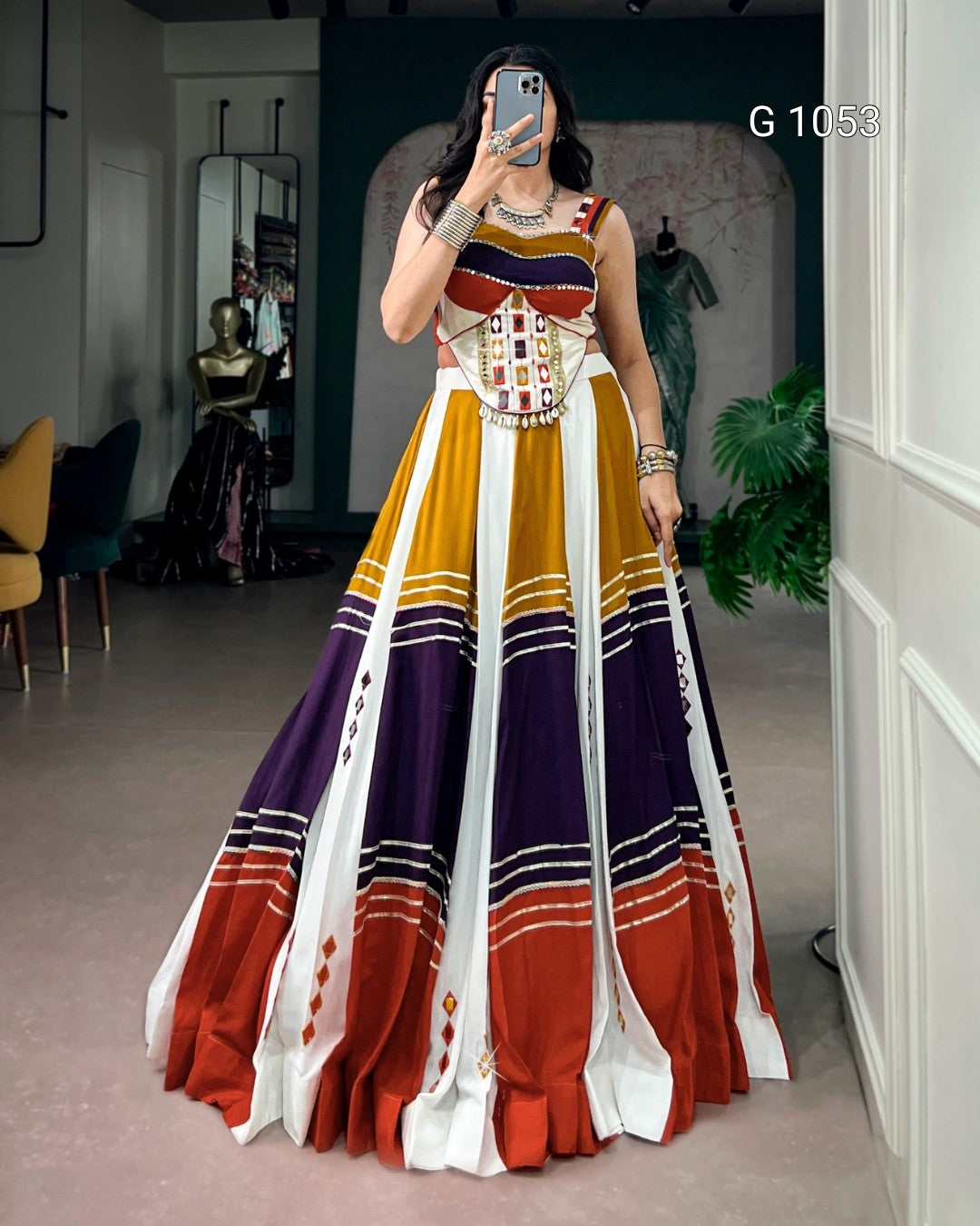 A woman takes a mirror selfie wearing a sleeveless, floor-length A-line gown with a geometric multicolor pattern of yellow, purple, orange, and white. The dress features a fitted bodice with a geometric print and a heavily embellished corset-style waistband with gold and silver beadwork.
