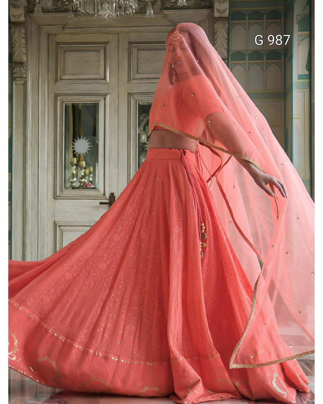 A woman modeling a peach-pink traditional Indian lehenga choli set with a matching sheer dupatta veil, featuring gold embroidery and border details, posed in front of an ornate wooden door. The code 'G 987' is visible in the top right corner.