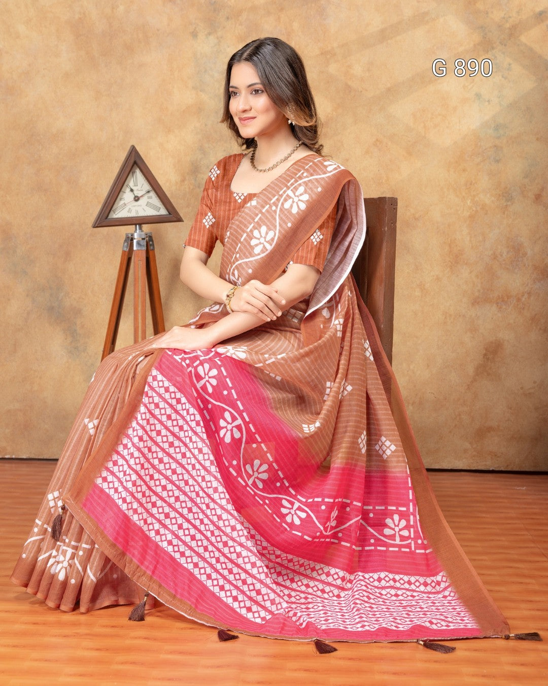 Woman wearing a traditional saree with a brown and red pattern, sitting on a chair against a beige wall.