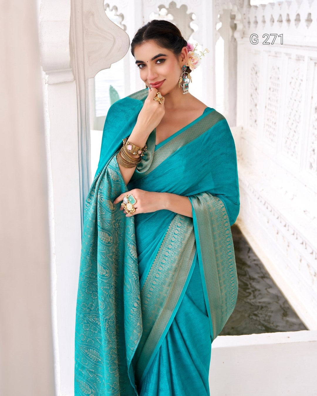 A woman wearing a teal blue saree with a metallic gold border, standing in a white architectural corridor. She is adorned with traditional jewelry and a flower in her hair.