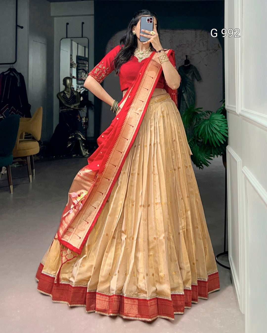A woman takes a mirror selfie wearing a traditional Indian outfit consisting of a beige flared skirt, a red blouse, and a red dupatta with gold zari borders. The setting appears to be a boutique or studio interior with clothing racks and plants in the background.