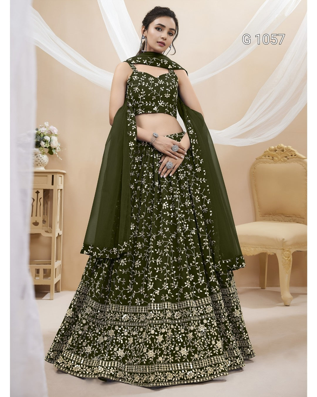 A woman models a dark olive green traditional Indian lehenga choli set with intricate gold and silver floral embroidery, set against a warm beige background with draped white fabric and a classic chair.