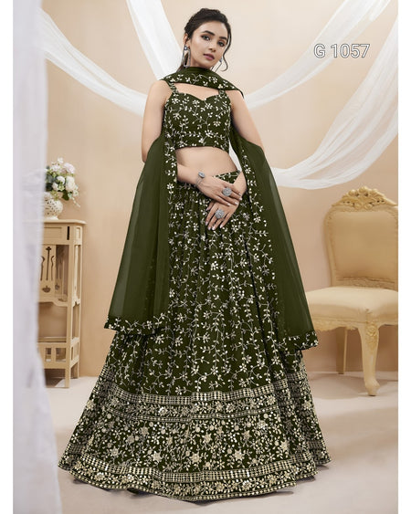A woman models a dark olive green traditional Indian lehenga choli set with intricate gold and silver floral embroidery, set against a warm beige background with draped white fabric and a classic chair.