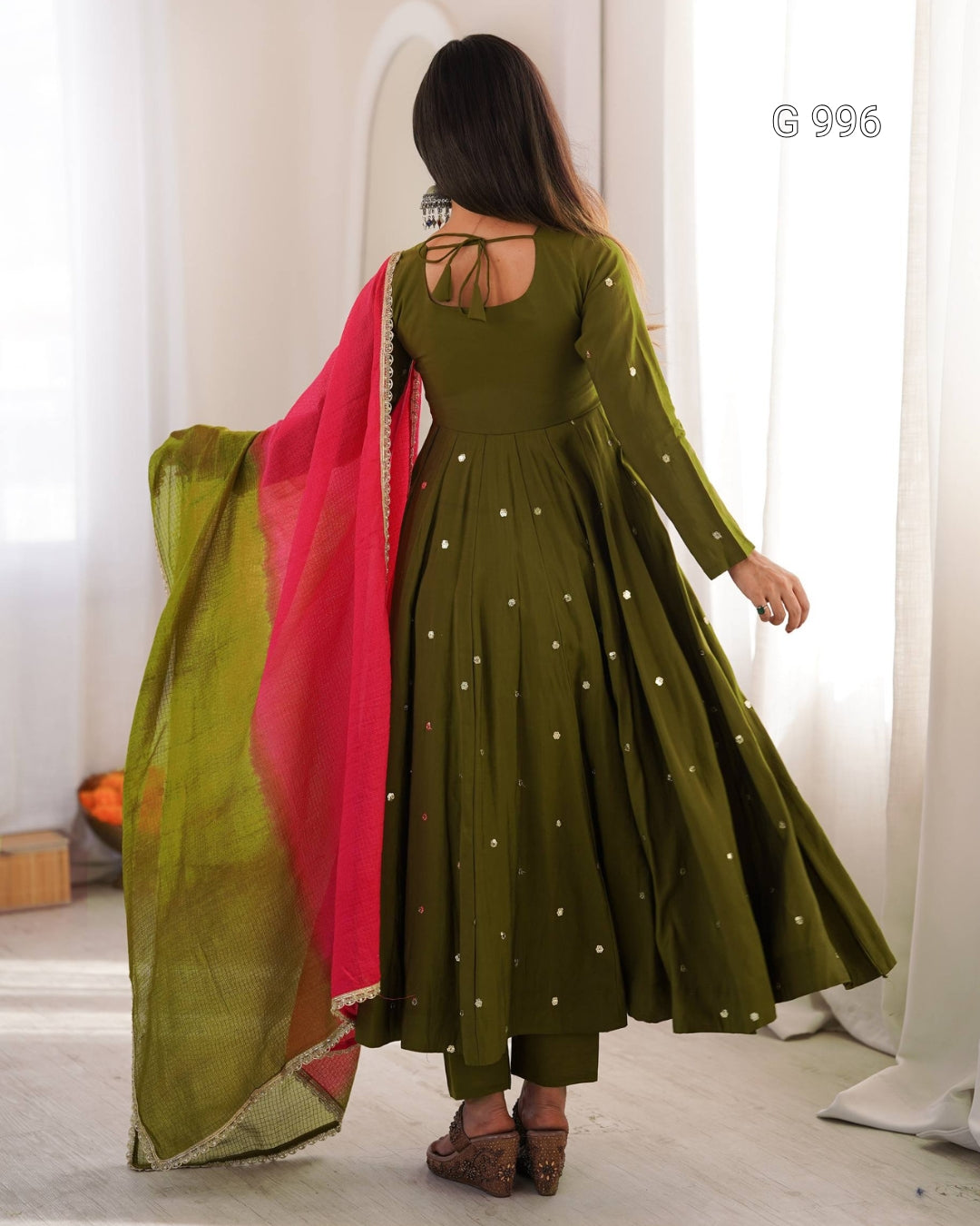 A woman shown from the back wearing an olive green long-sleeve Anarkali-style dress with small gold dot embellishments, paired with a bright pink dupatta draped over her arm, in a softly lit indoor setting.