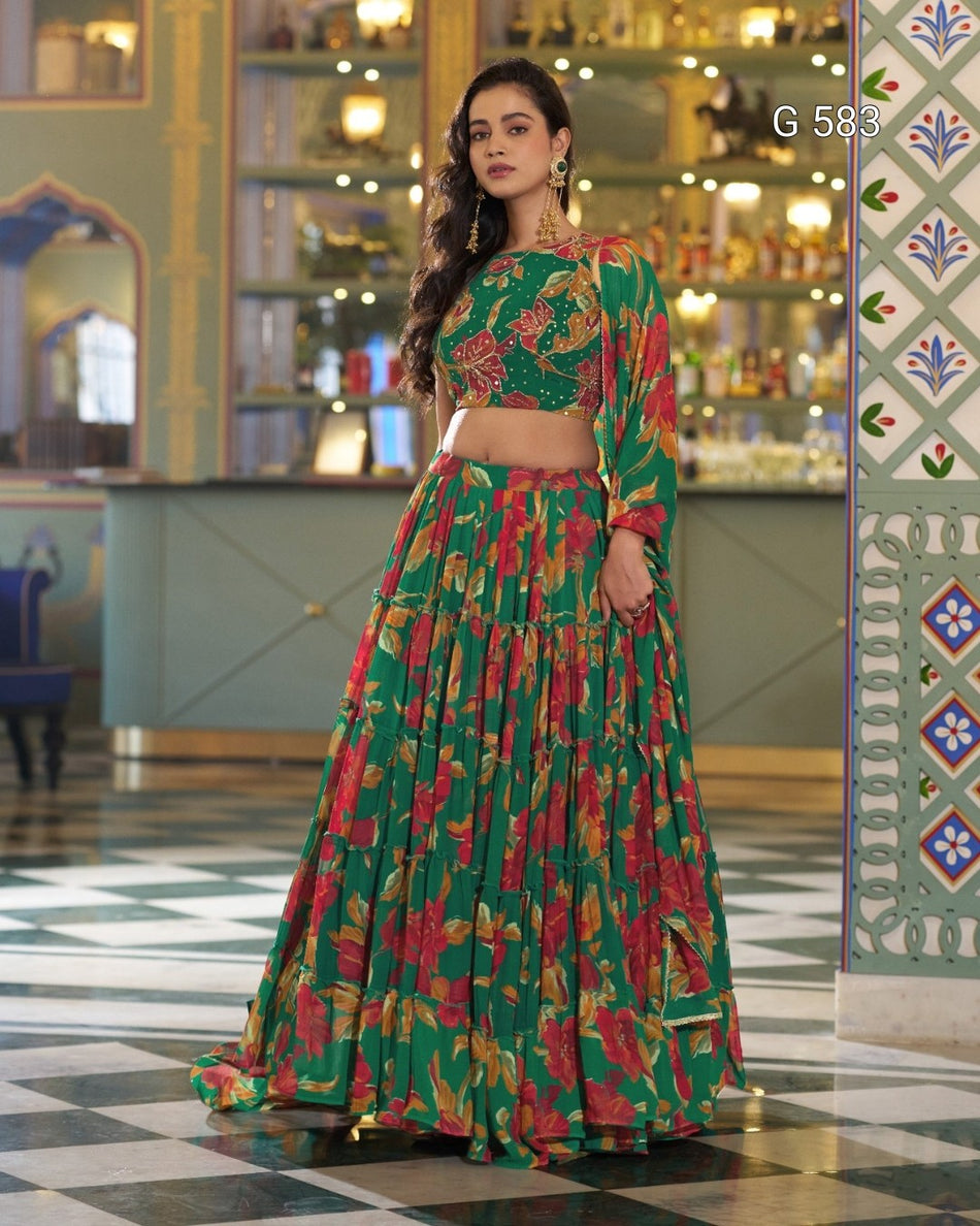 A woman models a green three-piece floral lehenga choli set in an ornate indoor setting. The set features a green long-sleeve crop top with red and gold floral embroidery, a matching green tiered skirt, and a coordinating green dupatta.