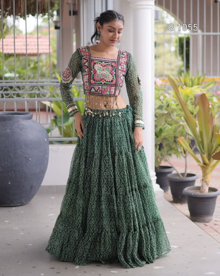 A woman models a traditional Indian ethnic outfit consisting of a green patterned long skirt (lehenga) and a matching embroidered green blouse with a colorful floral border, set against a porch background with potted plants.