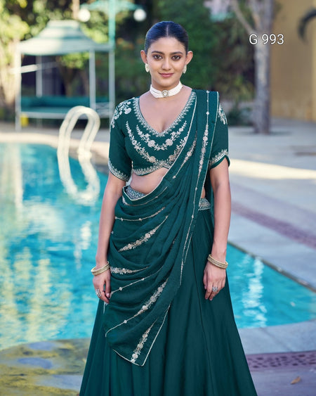 A woman stands by a swimming pool wearing a dark green traditional outfit with silver embroidery. The outfit consists of a sleeveless embroidered blouse and a matching draped skirt.