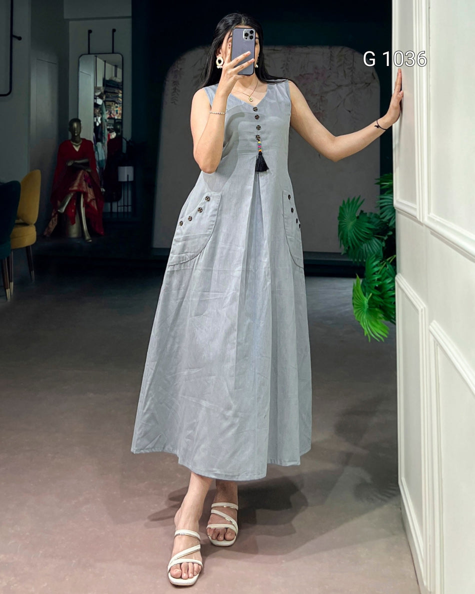 A woman takes a mirror selfie wearing a sleeveless light blue linen midi dress with a tassel tie at the neckline and decorative buttons on the pockets. The background is a boutique interior with a mannequin and a white wall panel.