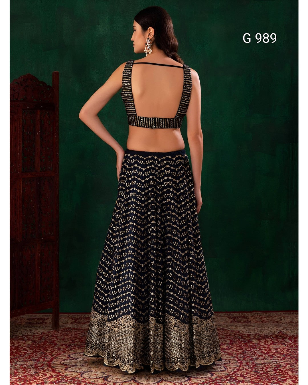 A woman shown from the back wearing a black and silver patterned lehenga choli set against a dark green textured backdrop. The set features a sleeveless backless crop top with embellished straps and a matching flared skirt with a metallic gold border at the hem.