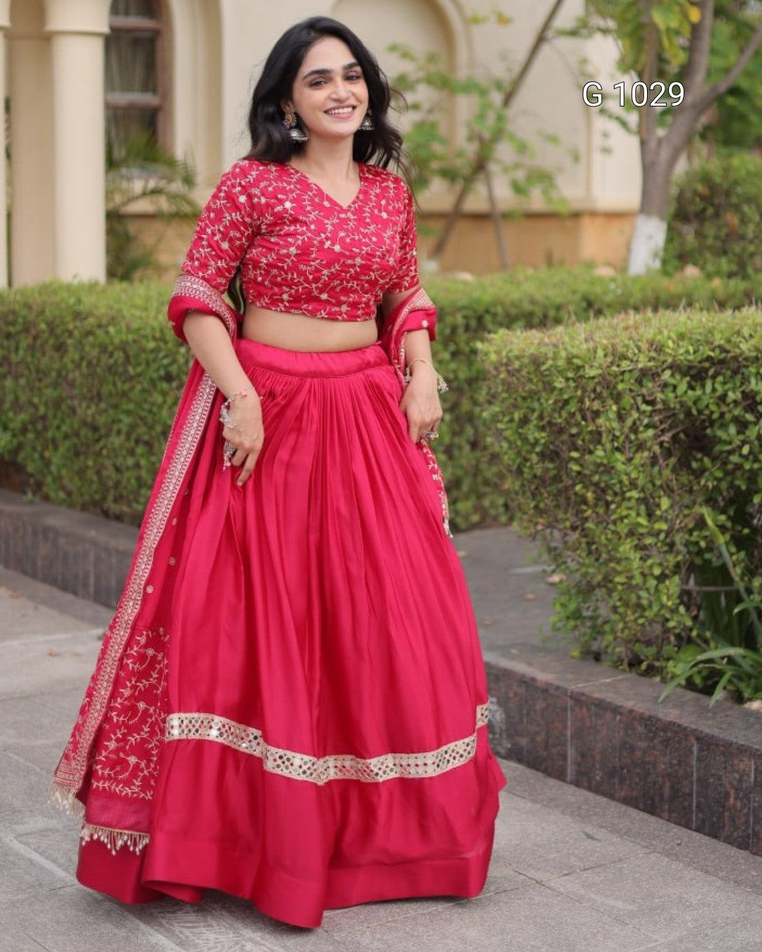A woman modeling a bright pink-red traditional Indian outfit with a heavily embroidered matching blouse, a full flared skirt, and a draped dupatta, standing outdoors in front of trimmed hedges and a building.