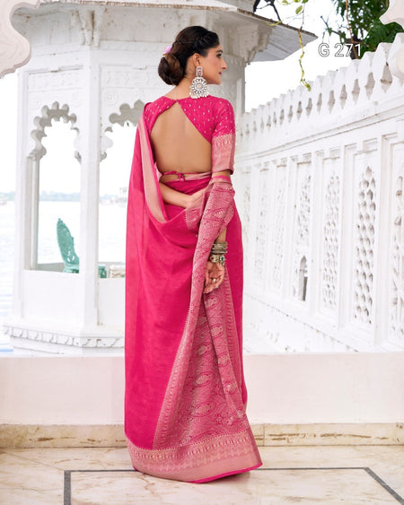 A woman wearing a bright pink saree with a matching blouse, photographed from the back in a white architectural setting with carved walls and a tiled floor.