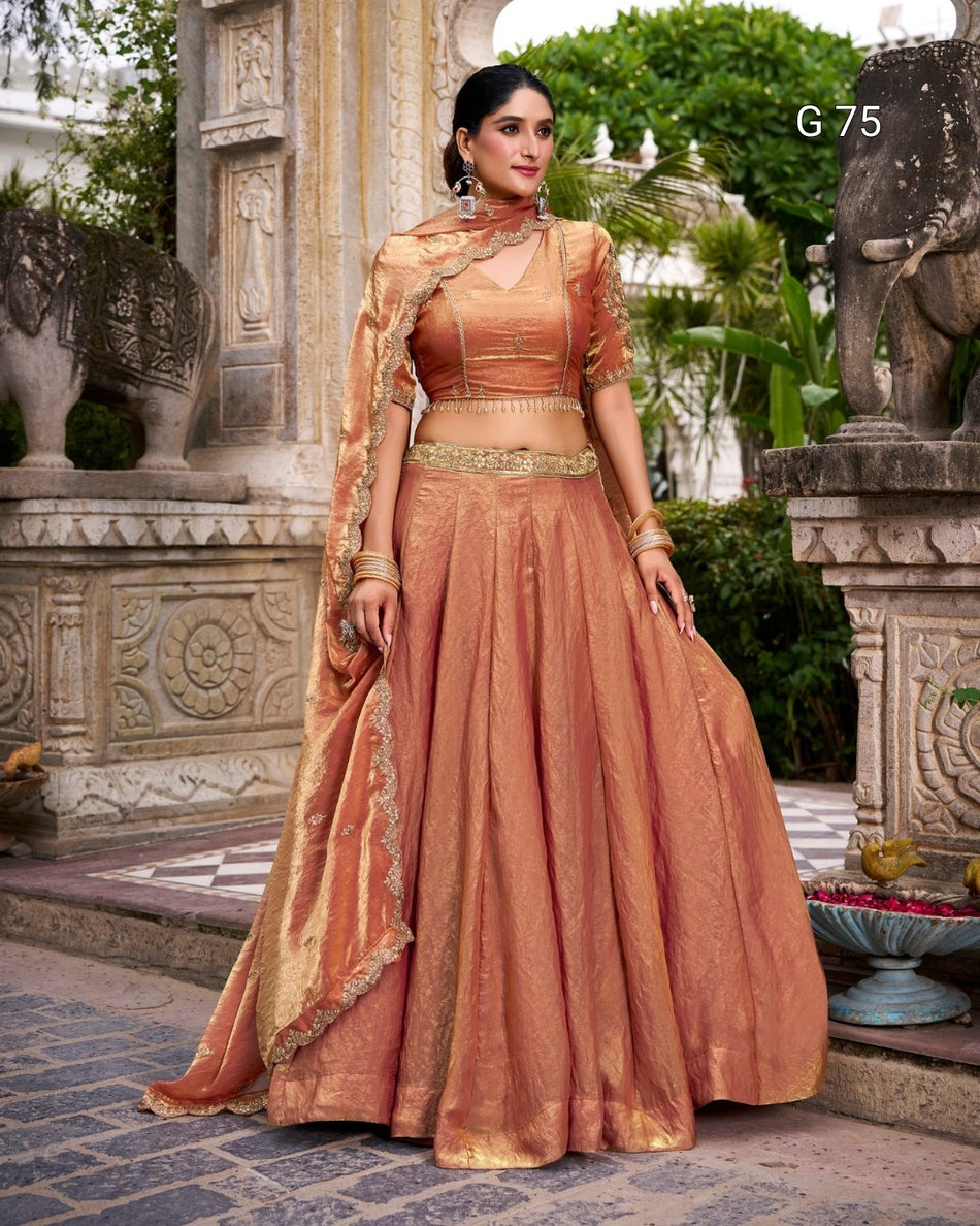 A woman models a traditional Indian salwar kameez outfit in a stone courtyard. The outfit consists of a peach-colored flared skirt, a matching embroidered blouse, and a draped dupatta.