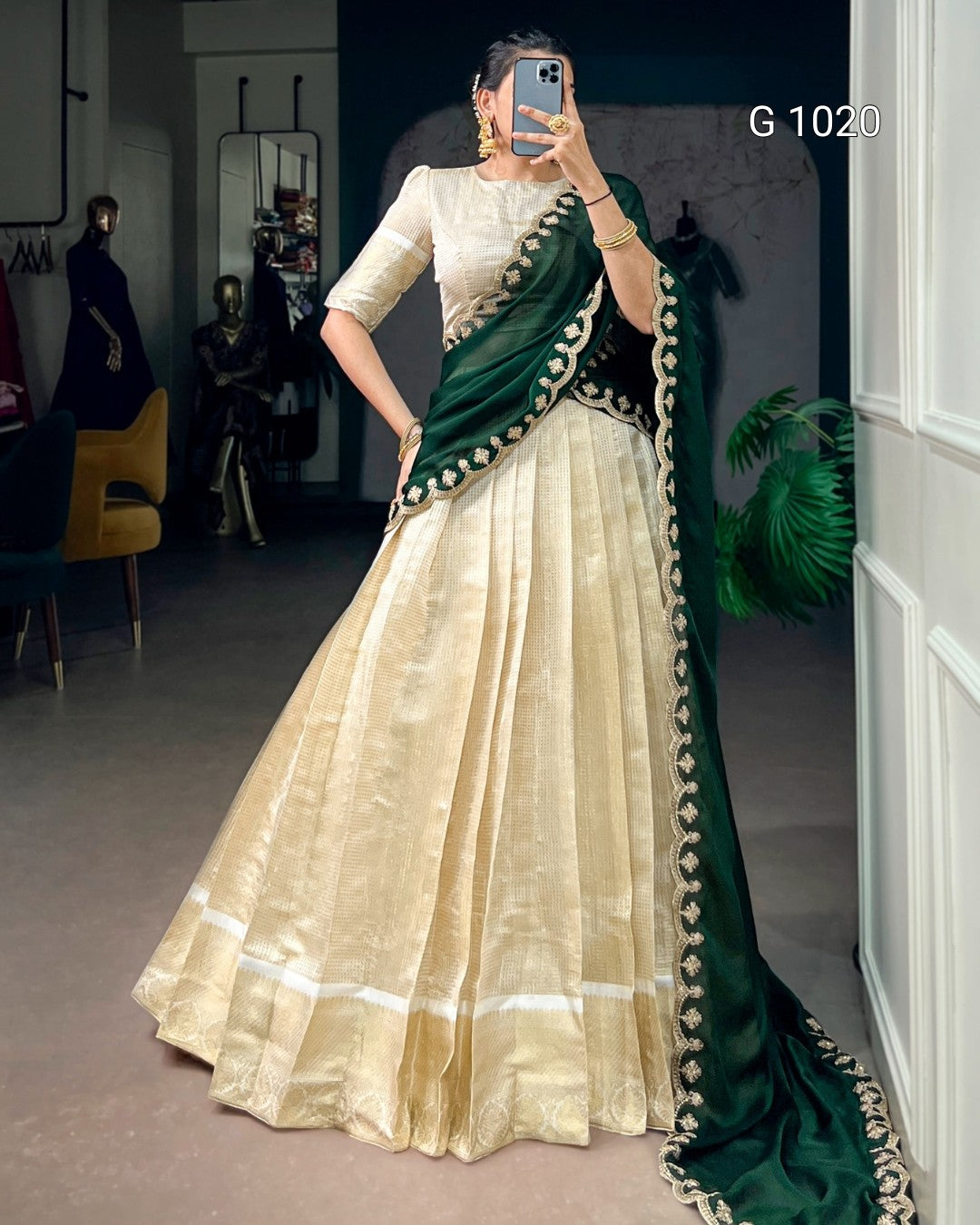 A woman takes a mirror selfie in a boutique wearing a traditional Indian lehenga choli. The outfit features a cream-colored pleated skirt (lehenga) and a matching cream blouse with a dark green border, draped with a dark green dupatta that has gold embroidery.