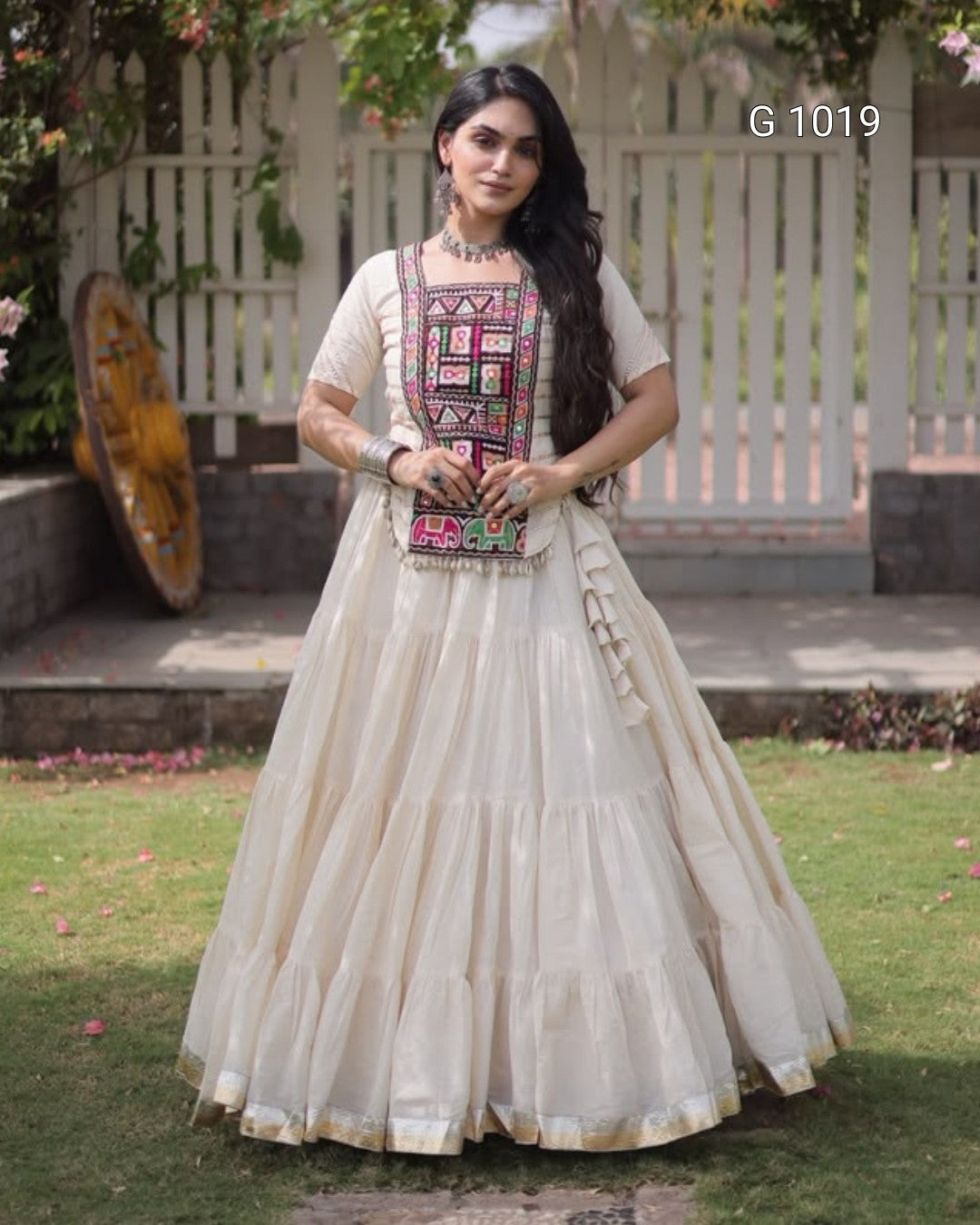 A woman stands outdoors in a garden wearing a cream-colored traditional Anarkali gown with a heavily embroidered multicolor bodice panel. She is holding a matching embroidered clutch bag.