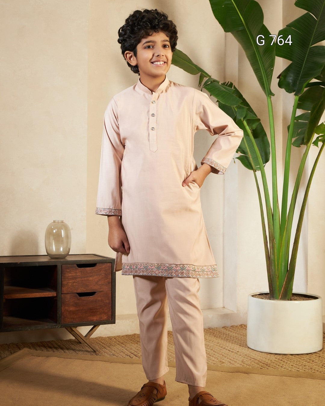Young boy in a light pink traditional outfit standing in a room with a plant and wooden cabinet.