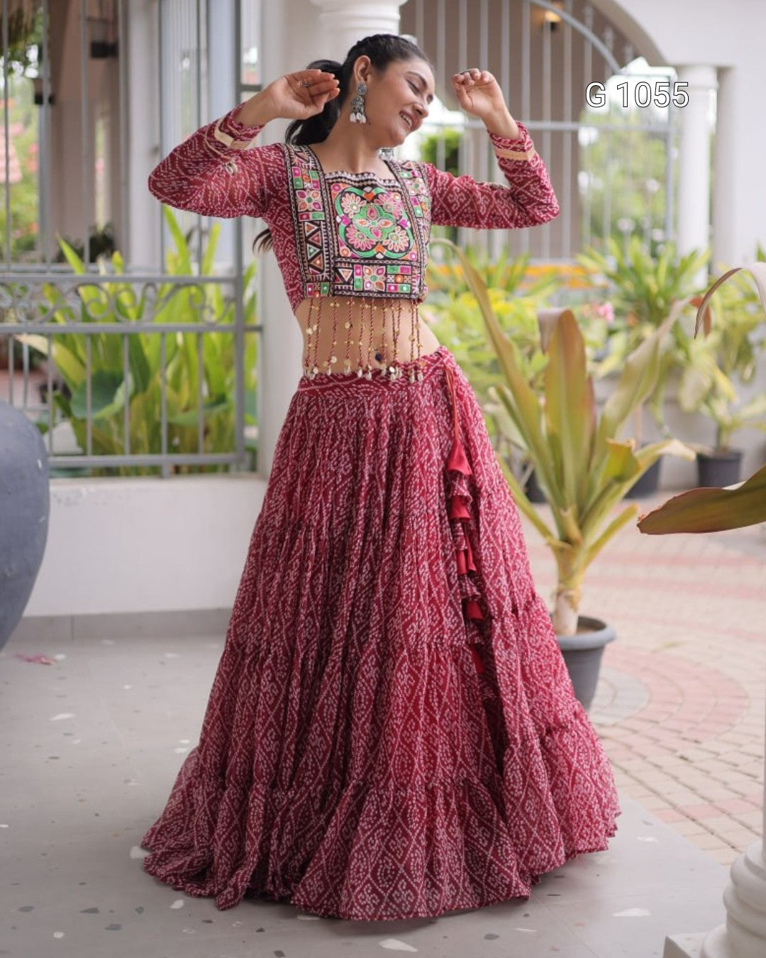 A woman modeling a traditional Indian lehenga choli dress in a dusty rose pink color with intricate multicolor embroidery on the bodice, set in an outdoor patio environment with plants.