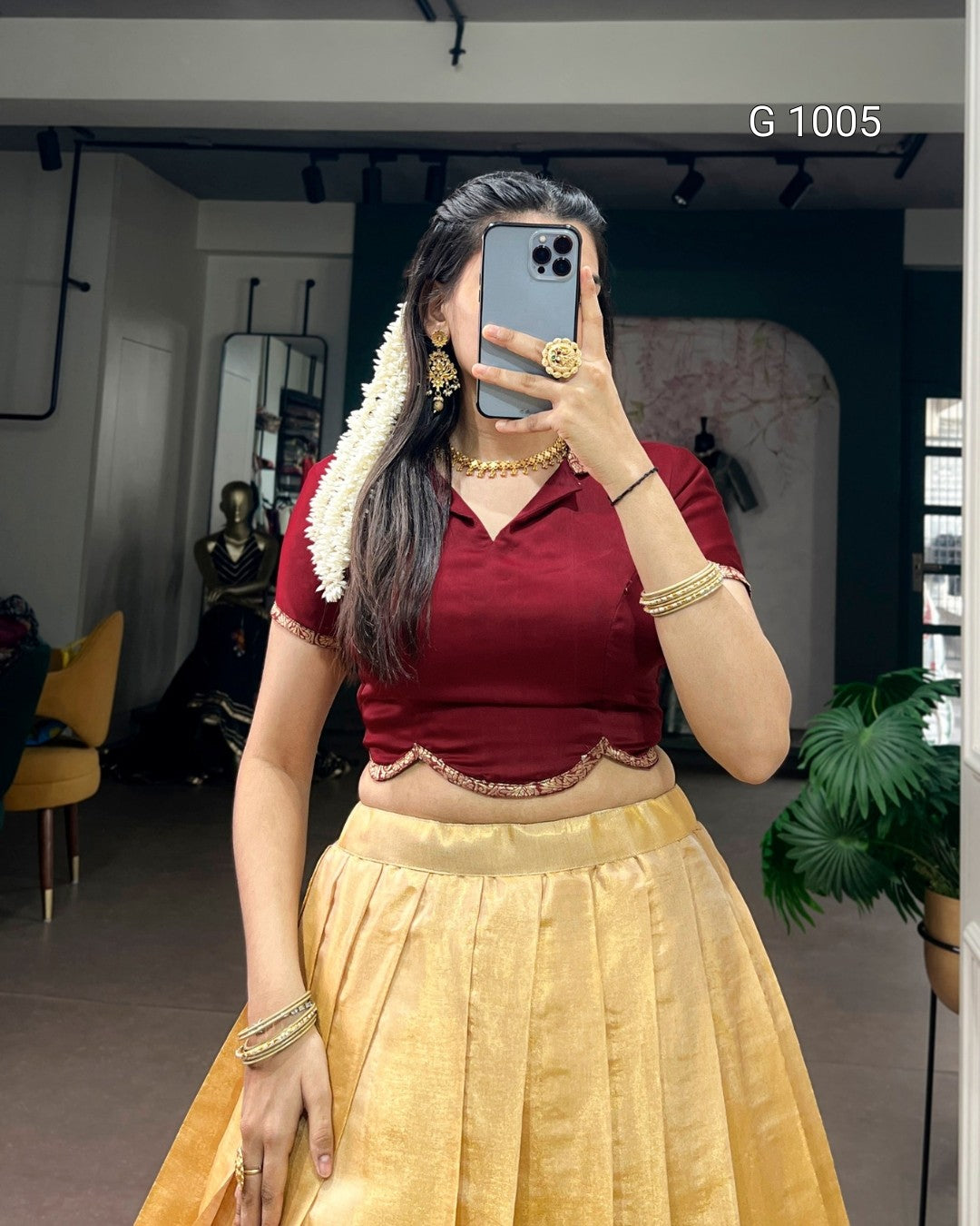 A woman takes a mirror selfie wearing a traditional Indian outfit consisting of a deep red short-sleeve blouse with a scalloped hem and a pleated gold skirt. She is accessorized with gold jewelry, including a necklace, bangles, and earrings, and a white floral garland is tied to her hair.