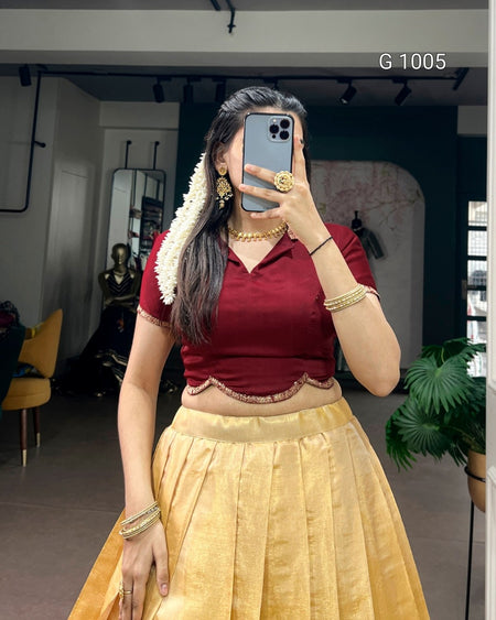 A woman takes a mirror selfie wearing a traditional Indian outfit consisting of a deep red short-sleeve blouse with a scalloped hem and a pleated gold skirt. She is accessorized with gold jewelry, including a necklace, bangles, and earrings, and a white floral garland is tied to her hair.