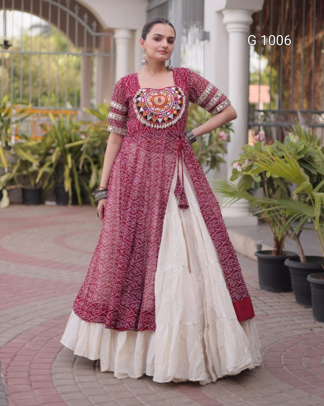 A woman modeling a traditional Indian salwar suit in a courtyard setting. The outfit features a dusty rose printed kurta with a heavily embroidered circular yoke, paired with a cream-colored flared skirt.