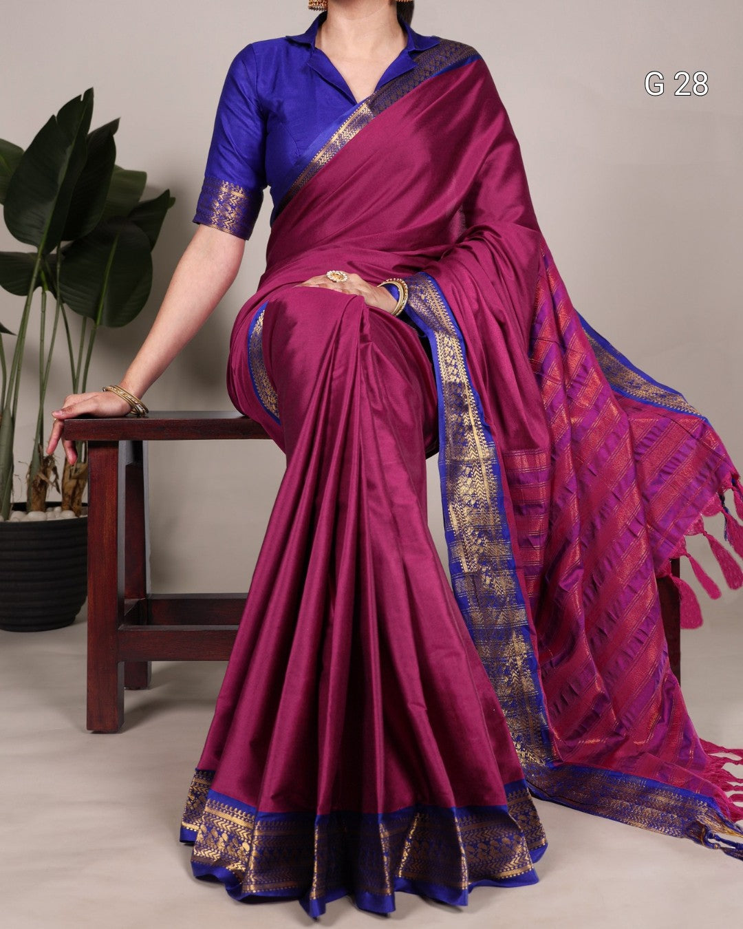 Woman wearing a purple saree with a blue border, sitting on a wooden stool.