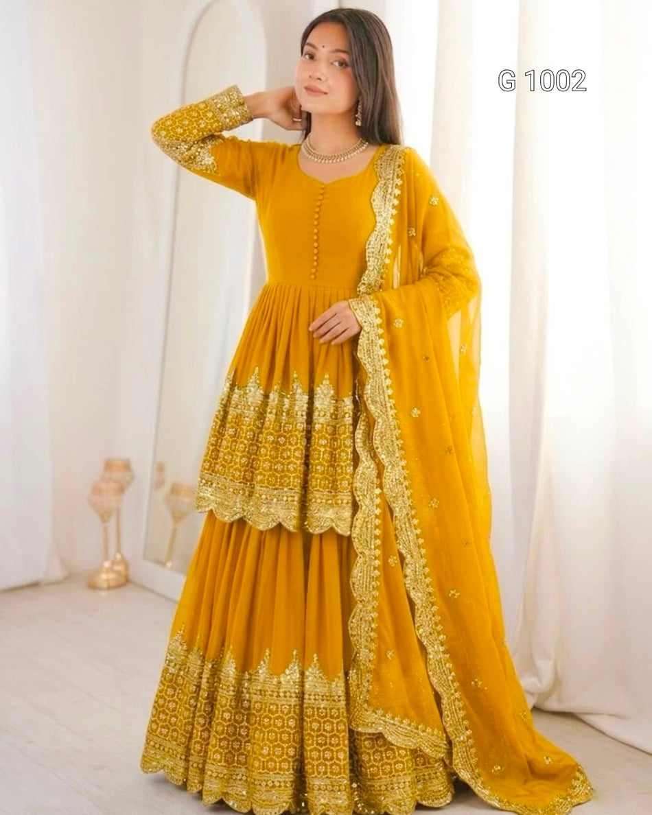 A woman models a traditional Indian outfit consisting of a mustard yellow Anarkali gown with intricate gold embroidery, matching embroidered dupatta, and a long skirt. The background is a soft, warm-toned indoor setting with white curtains.