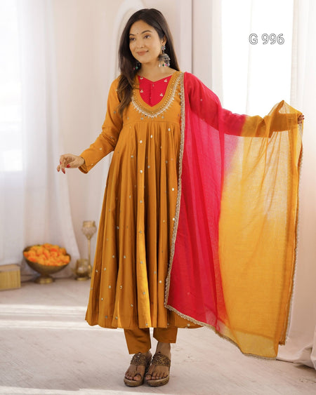 A woman models a traditional Indian outfit consisting of a mustard yellow Anarkali kurta with a red embroidered neckline, matching mustard pants, and a red-to-yellow gradient dupatta, set against a bright, softly lit indoor background.