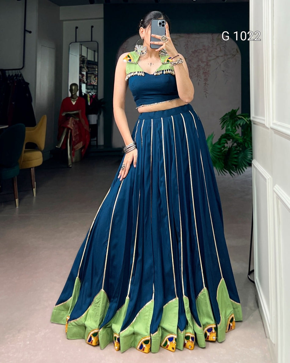 A woman takes a mirror selfie in a boutique wearing a dark teal blue traditional Indian outfit with a sleeveless crop top, a flared skirt with vertical gold stripes, and a green and gold peacock-inspired border at the hem. The text 'G 1022' is visible in the top right corner.