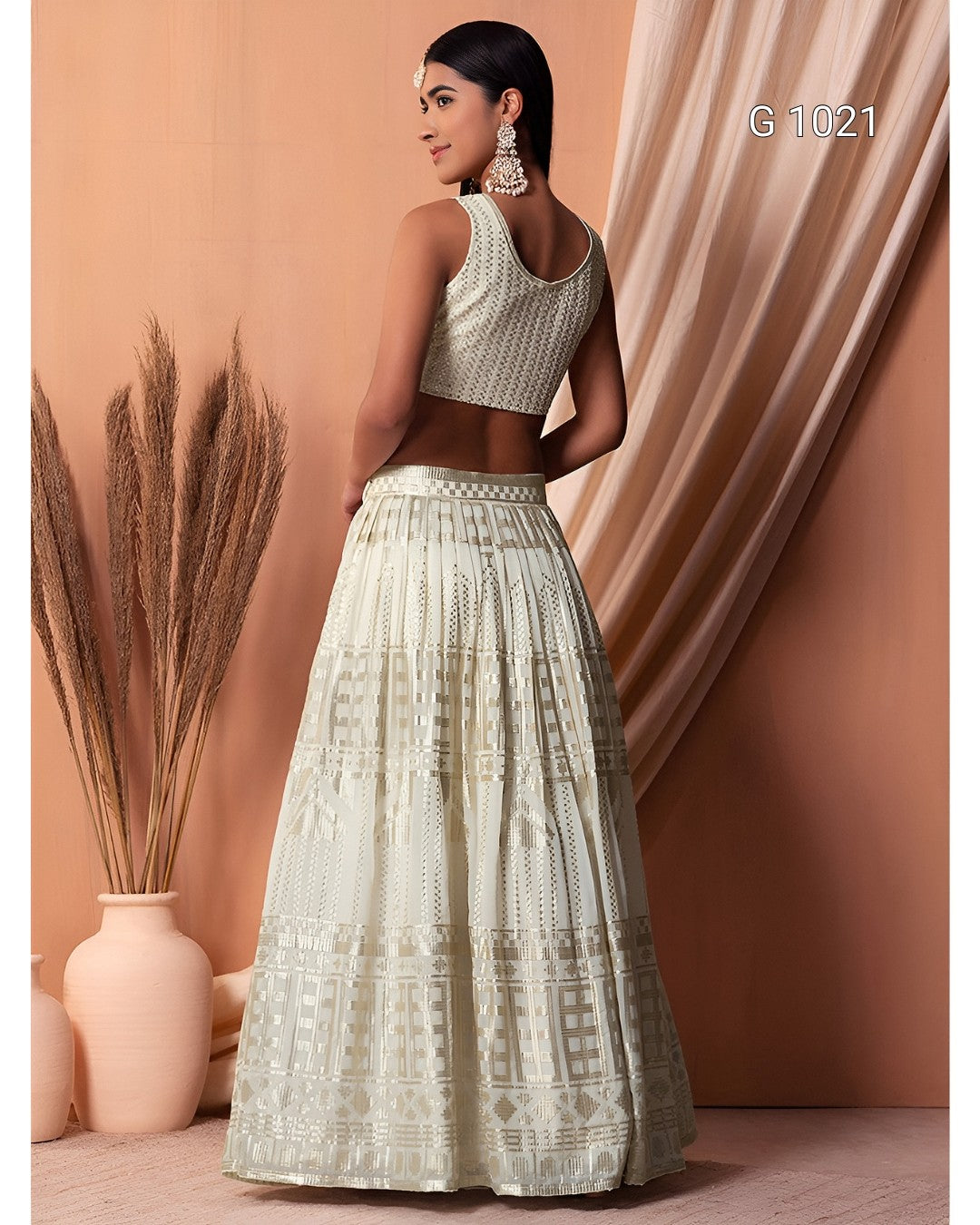A woman modeling a light beige embroidered traditional outfit with a sleeveless embellished top and a full skirt, posed against a warm peach backdrop with draped fabric and dried pampas grass.