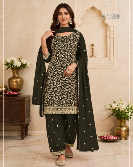 A woman models a dark green three-piece salwar kameez suit with intricate gold and silver floral embroidery, paired with a matching dupatta, set against a traditional beige architectural background.