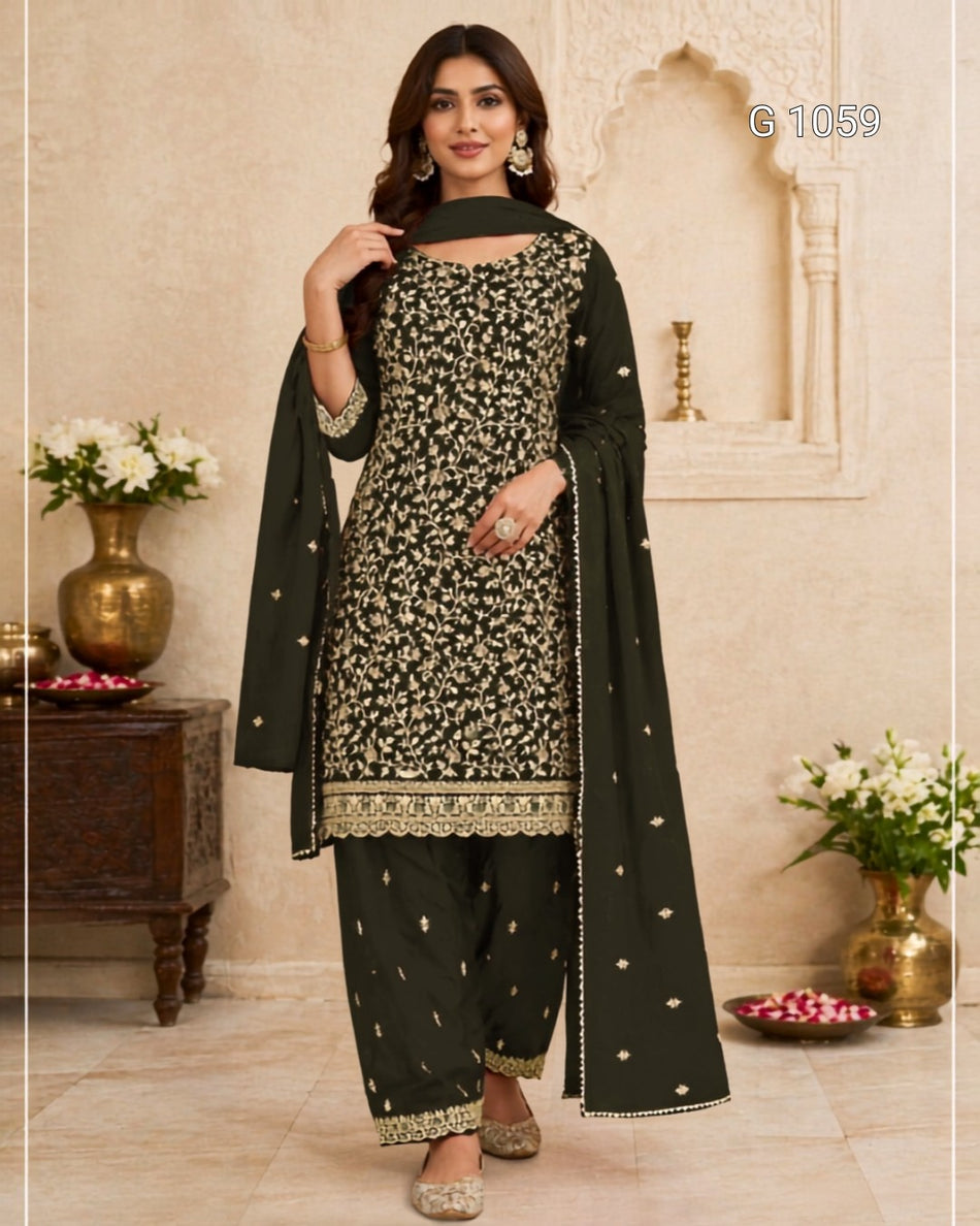 A woman models a dark green three-piece salwar kameez suit with intricate gold and silver floral embroidery, paired with a matching dupatta, set against a traditional beige architectural background.