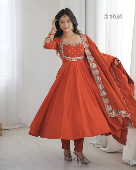 A woman models a traditional Indian outfit consisting of a rust-orange Anarkali gown with intricate silver embroidery, matching pants, and a draped dupatta. The background is a bright, minimalist room with white curtains.