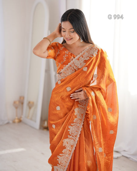 A woman modeling an orange saree with intricate white floral embroidery along the border and blouse, standing in a bright, minimalist indoor setting.