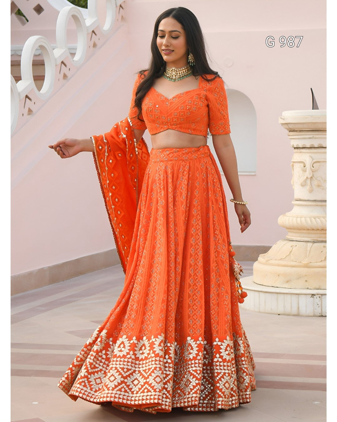 An orange embroidered Indian ethnic wear set consisting of a flared skirt, a matching crop top, and a dupatta, modeled by a woman in front of a light pink architectural background.