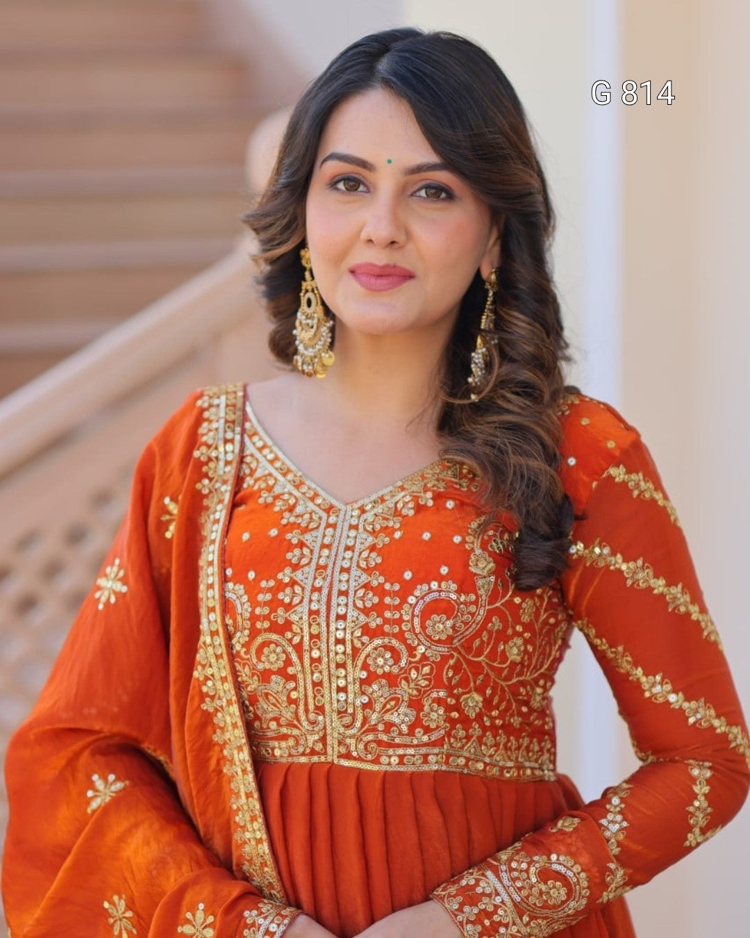 Woman wearing an orange traditional outfit with gold embroidery indoors.