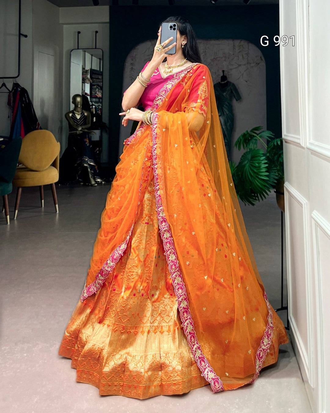 A woman wearing a traditional Indian saree in orange and pink, taking a mirror selfie in an indoor boutique setting. The saree features a bright orange body with a pink and gold border, and a heavily embroidered golden-yellow zari border.