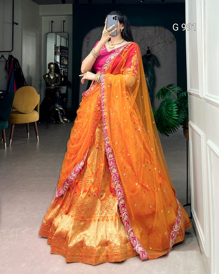 A woman wearing a traditional Indian saree in orange and pink, taking a mirror selfie in an indoor boutique setting. The saree features a bright orange body with a pink and gold border, and a heavily embroidered golden-yellow zari border.
