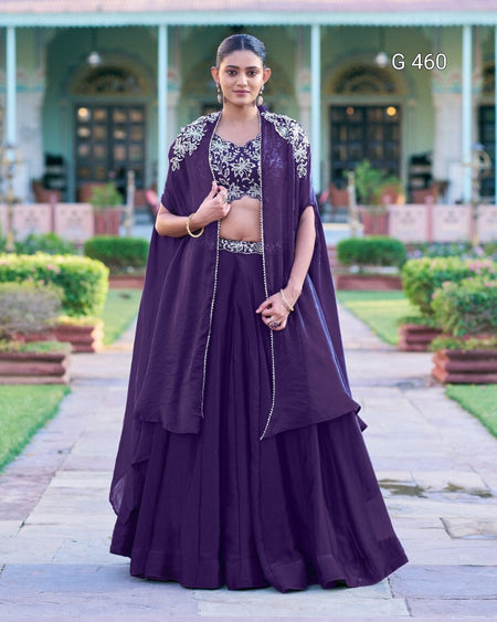 A woman stands outdoors in a formal garden courtyard wearing a dark purple traditional outfit with a matching draped outer layer, embellished silver embroidery on the chest, and a long flared skirt. The background shows a symmetrical building with columns and greenery.