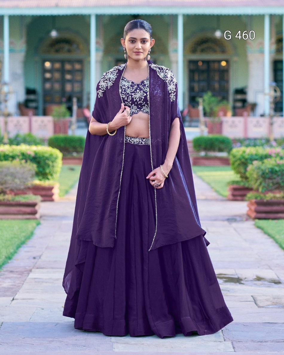 A woman stands outdoors in a formal garden courtyard wearing a dark purple traditional outfit with a matching draped outer layer, embellished silver embroidery on the chest, and a long flared skirt. The background shows a symmetrical building with columns and greenery.