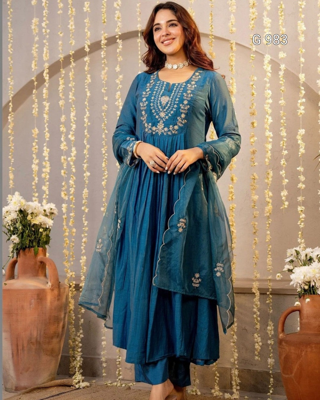 A woman models a teal blue ethnic salwar kameez suit with intricate silver embroidery on the chest, paired with a matching sheer dupatta, set against an arched backdrop with hanging floral garlands.